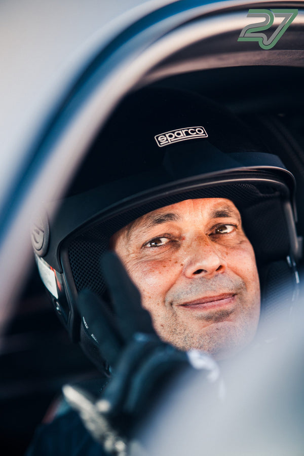 Älterer Fahrer mit schwarzem Sparco-Helm blickt zufrieden lächelnd zur Seite im Auto