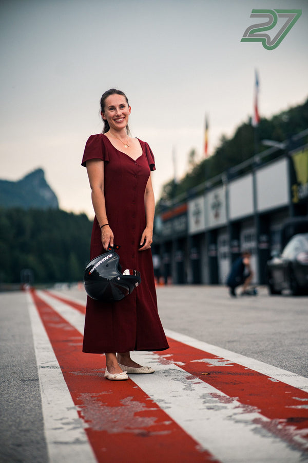 Frau im weinroten Kleid steht mit Helm in der Boxengasse, dahinter die Berge des Salzburgrings