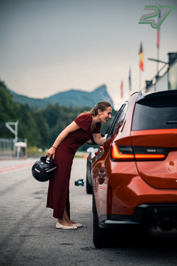 Die Frau im weinroten Kleid beugt sich lachend ins Fenster des orangen BMW, Berge dahinter