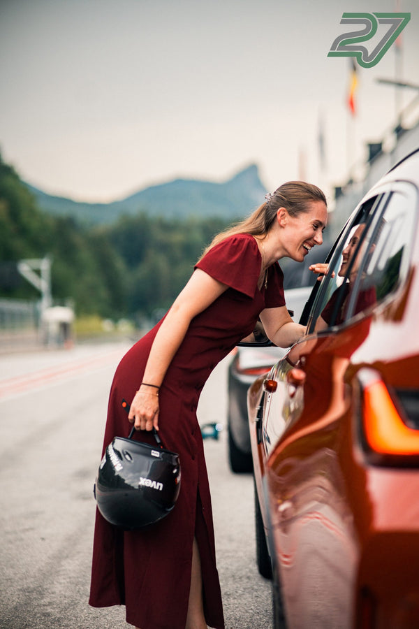 Aus der Nähe lacht die Frau im weinroten Kleid durch das offene Fenster des orangen BMW