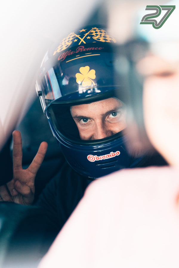 Nahaufnahme eines Fahrers im dunkelblauen Alfa-Romeo-Helm, der das Peace-Zeichen in die Kamera hält