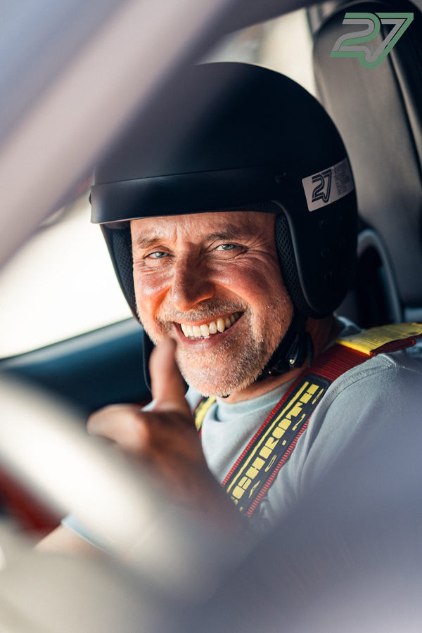 Lachender älterer Fahrer mit schwarzem 27-Helm und grauem Overall zeigt den Daumen hoch