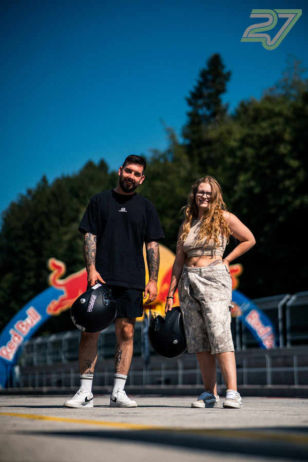 Ein Mann und eine Frau stehen mit Helmen in der Hand vor dem Red-Bull-Torbogen im Sonnenlicht