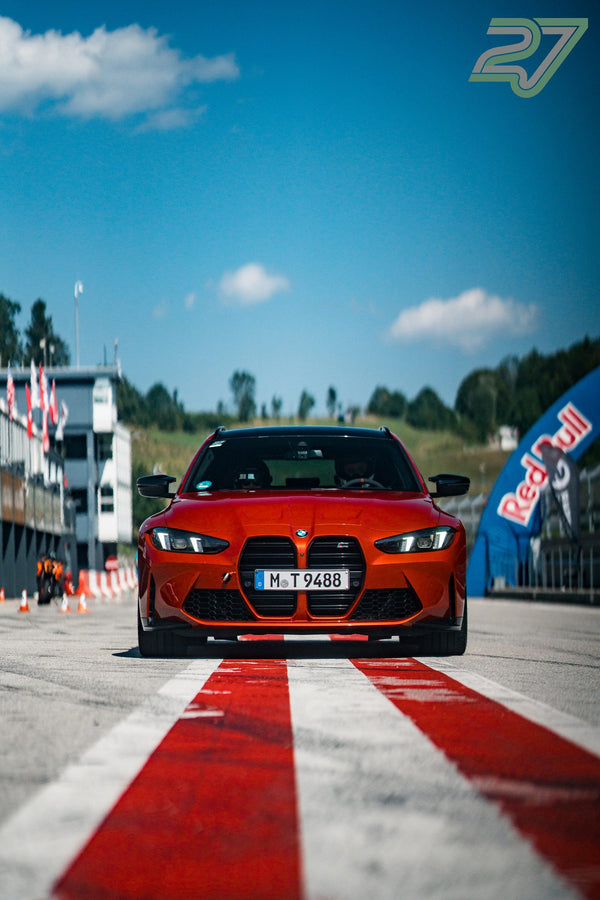 Oranger BMW M3 fährt geradeaus auf die Kamera zu, rechts flattert eine Red-Bull-Fahne