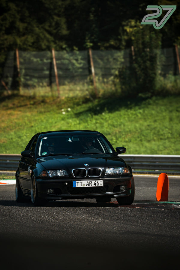 Schwarzer BMW 3er rollt durch die Schattenkurve an einem orangen Pylon vorbei, Beifahrer mit Kappe