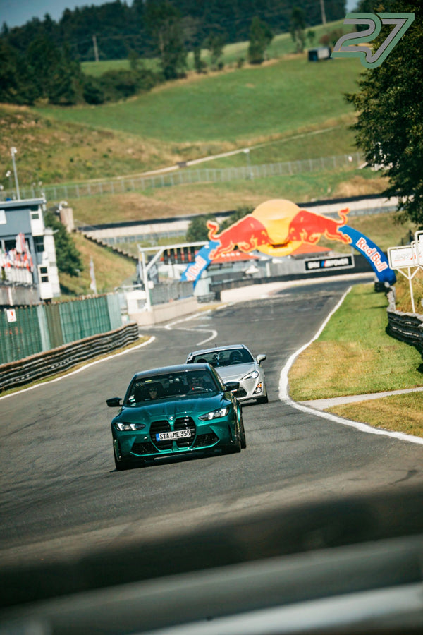 Dunkelgrüner BMW M3 rollt bergab auf den Red-Bull-Bogen zu, dahinter ein silberner Toyota