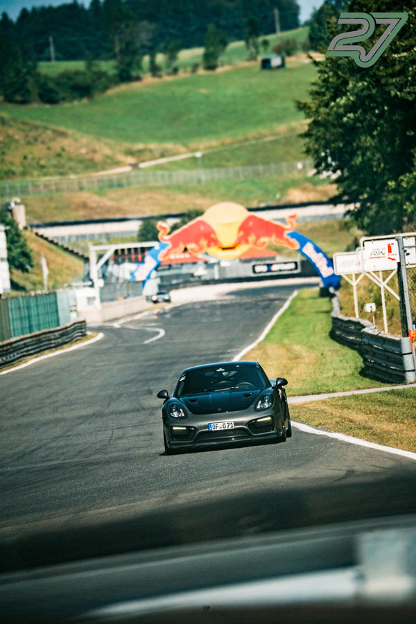Anthrazitfarbener Porsche Cayman rollt durch die Kurve unterhalb des Red-Bull-Bogens