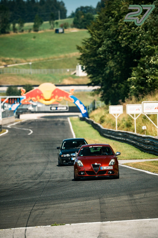 Roter Alfa Romeo Giulietta führt vor einem dunklen BMW auf den Red-Bull-Bogen zu