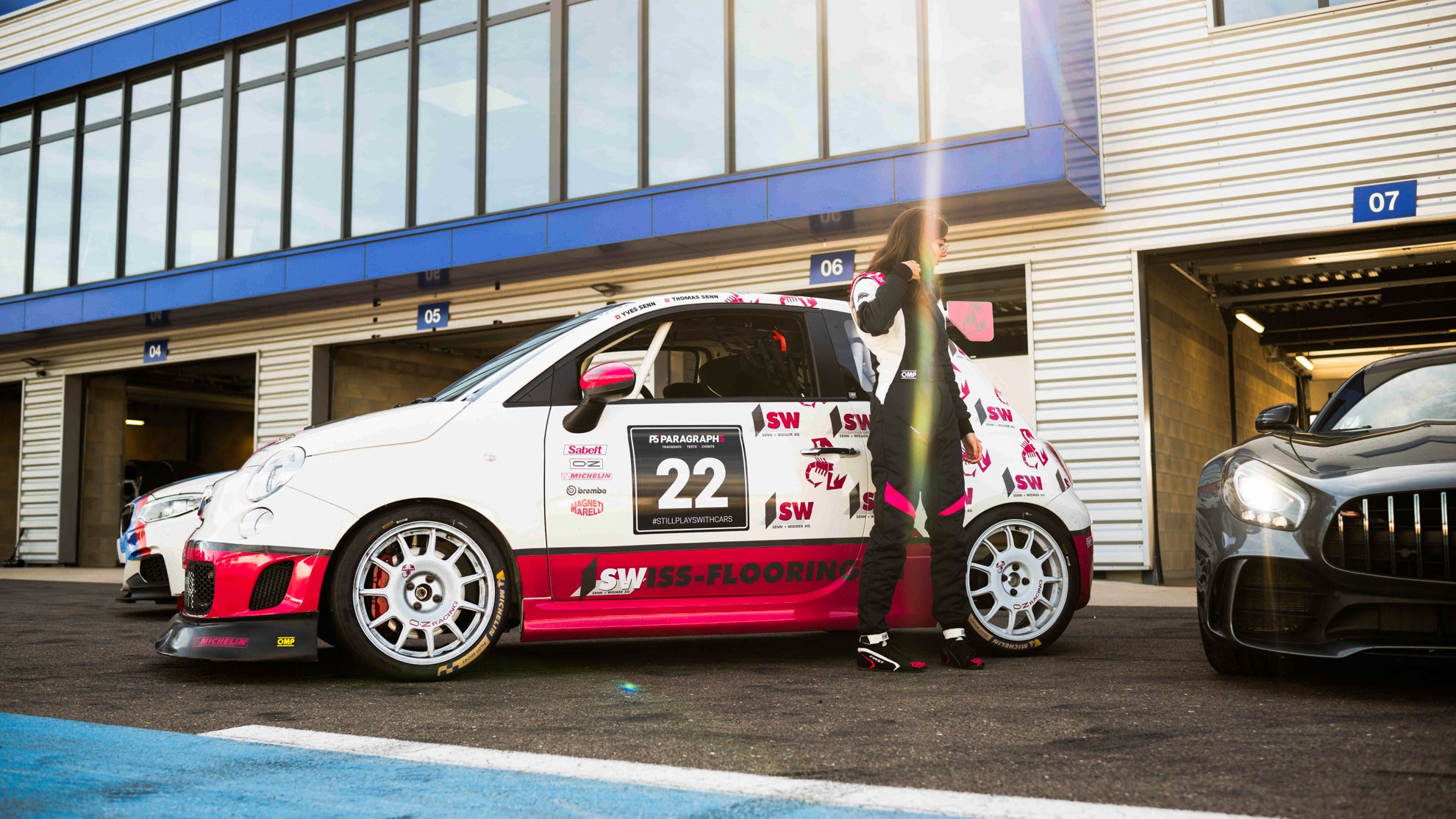 Weiß-pinker Abarth 595 Cup mit weißen OZ Felgen und seiner Fahrerin im OMP-Rennanzug neben einem AMG GTR auf dem Circuit Dijon-Prenois.